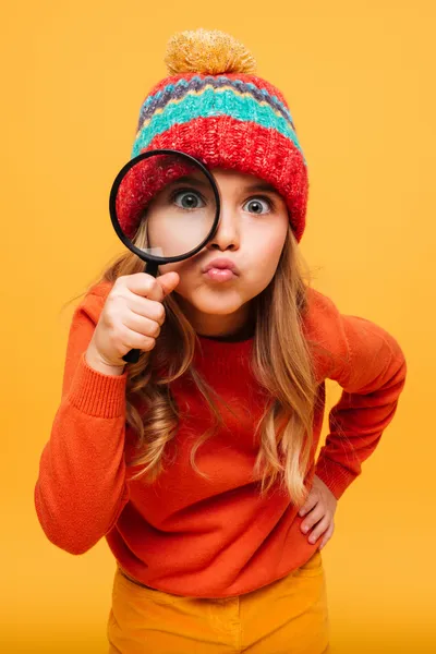 The image features a playful young girl holding a magnifying glass up to her eye, making one eye appear larger for a humorous effect. She's puffing her cheeks and pursing her lips in a funny, exaggerated expression. The girl is wearing a bright orange sweater and a colorful knitted beanie with a pom-pom on top, suggesting it might be cold. Her hair is long and appears to be a light color. The background is a warm, solid yellow, complementing her vibrant attire and adding to the playful nature of the photo.