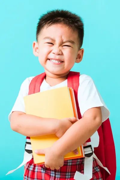 The image depicts a young boy with a spirited expression, tightly hugging a yellow folder to his chest. He is squinting his eyes and smiling broadly, which could indicate excitement or pride. The boy is dressed in a white t-shirt with a red backpack strapped to his shoulders, and he's wearing a plaid pair of shorts. The background is a bright turquoise blue, creating a cheerful and vibrant contrast to his outfit. This scene suggests a moment of joy, perhaps related to school or achievement.