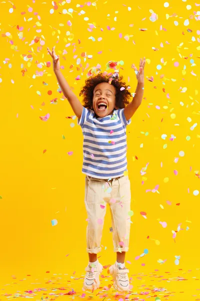 This is an image of a joyful child with arms raised exuberantly, surrounded by a shower of colorful confetti. The child is wearing a blue and white striped sleeveless top and light-colored cropped pants, complemented with white sneakers. The child's hair is styled in small, natural curls and is adorned with flowers. With a wide-open mouth in mid-laugh and eyes closed in delight, the child's expression is one of pure happiness. The background is a solid, warm yellow, enhancing the festive and vibrant atmosphere of the celebration captured in this moment.