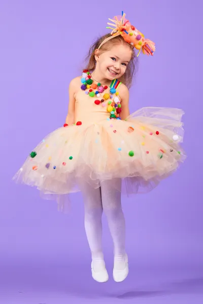The image features a young girl with a beaming smile, posing against a lavender backdrop. She is dressed in a whimsical costume that includes a tutu adorned with colorful pompoms, white tights, and ballet flats. Her blonde hair is styled in a playful manner, accessorized with a headpiece featuring more pompoms. Her attire and the joyous expression on her face give off a festive and playful vibe, as if she's dressed for a celebration or a dance performance.