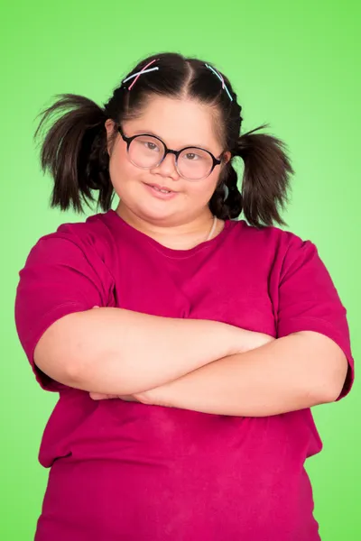 This is an image of a girl with a confident stance, wearing a solid dark pink t-shirt. She has her arms crossed and is smiling, giving a strong and assured impression. Her hair is styled in two high ponytails with what appears to be colorful strands wrapped around them. She is wearing round glasses and has a joyful expression on her face. The background is a bright, solid green, providing a stark contrast that highlights her figure and cheerful demeanor.