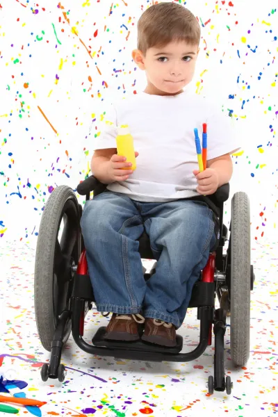 The image shows a young boy sitting in a wheelchair against a red background. He has short hair and is holding a yellow container, possibly of bubbles, in one hand and a pair of colorful markers in the other. He is wearing a white t-shirt, blue jeans, and brown shoes. His expression is calm and attentive, and he seems to be engaging with the viewer or ready to participate in an activity. The wheelchair suggests mobility support, and the child's readiness with art supplies indicates a setting for play or learning.